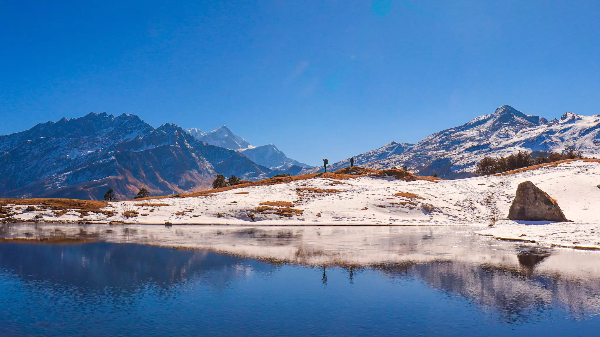 A lake in the foresground with a snowy trail and snow-capped mountains in the distance on a cloudless, sunny day.