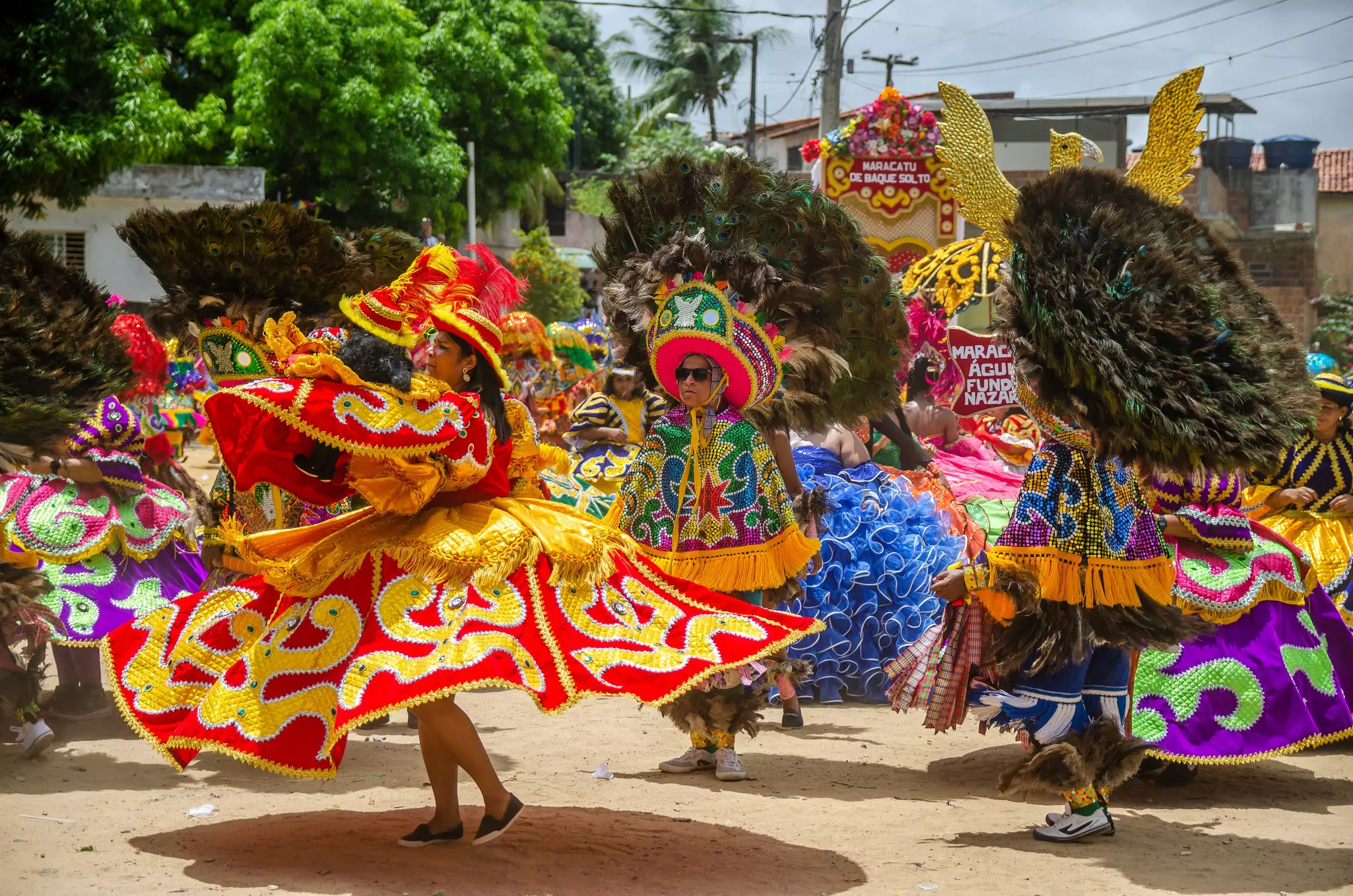 A woman wearing a yellow and gold patterned costume dances in the street. Nearby are people with vast feather-based headdresses.