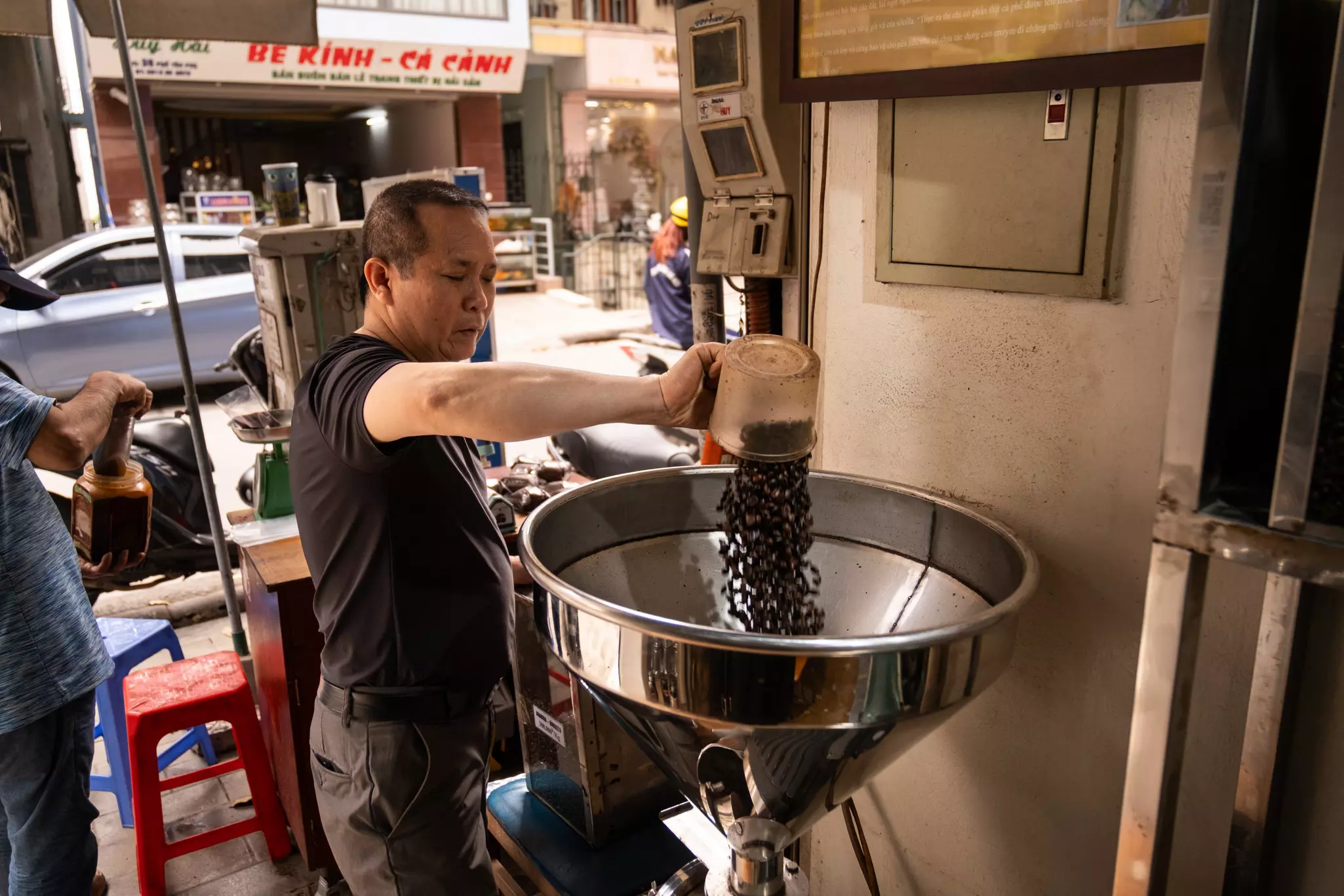 A man empties a bucket of coffee beans into a large grinder at a cafe