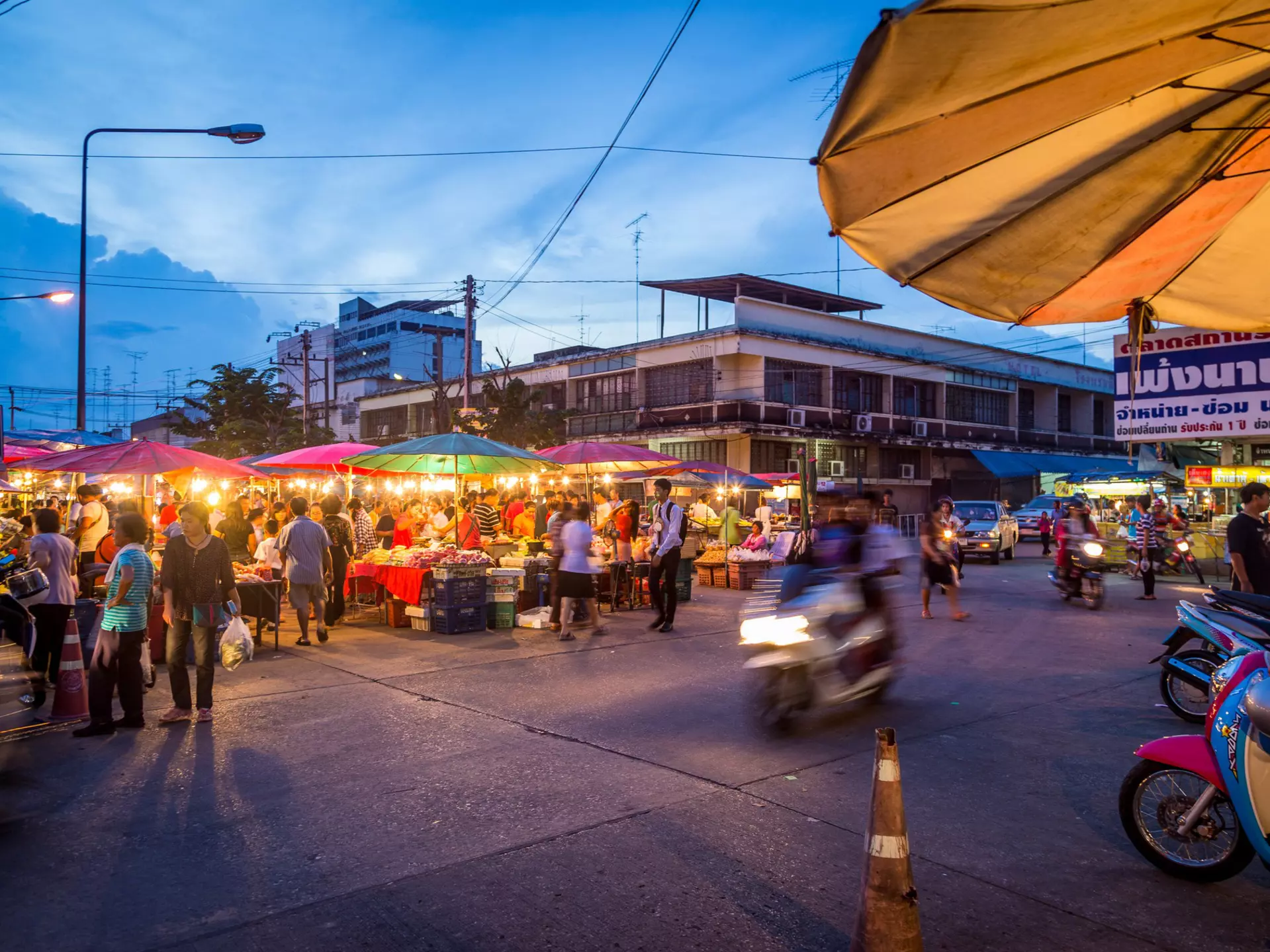 A blurred-out motorcycle drives past crowds at a night market on the streets of Phitsanulok, Thailand at dusk