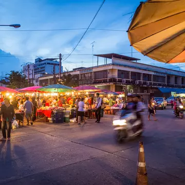 A blurred-out motorcycle drives past crowds at a night market on the streets of Phitsanulok, Thailand at dusk
