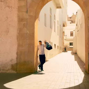 A man carrying a bag walks through a sunlit stone archway into a white-walled lane in Casablanca's old medina