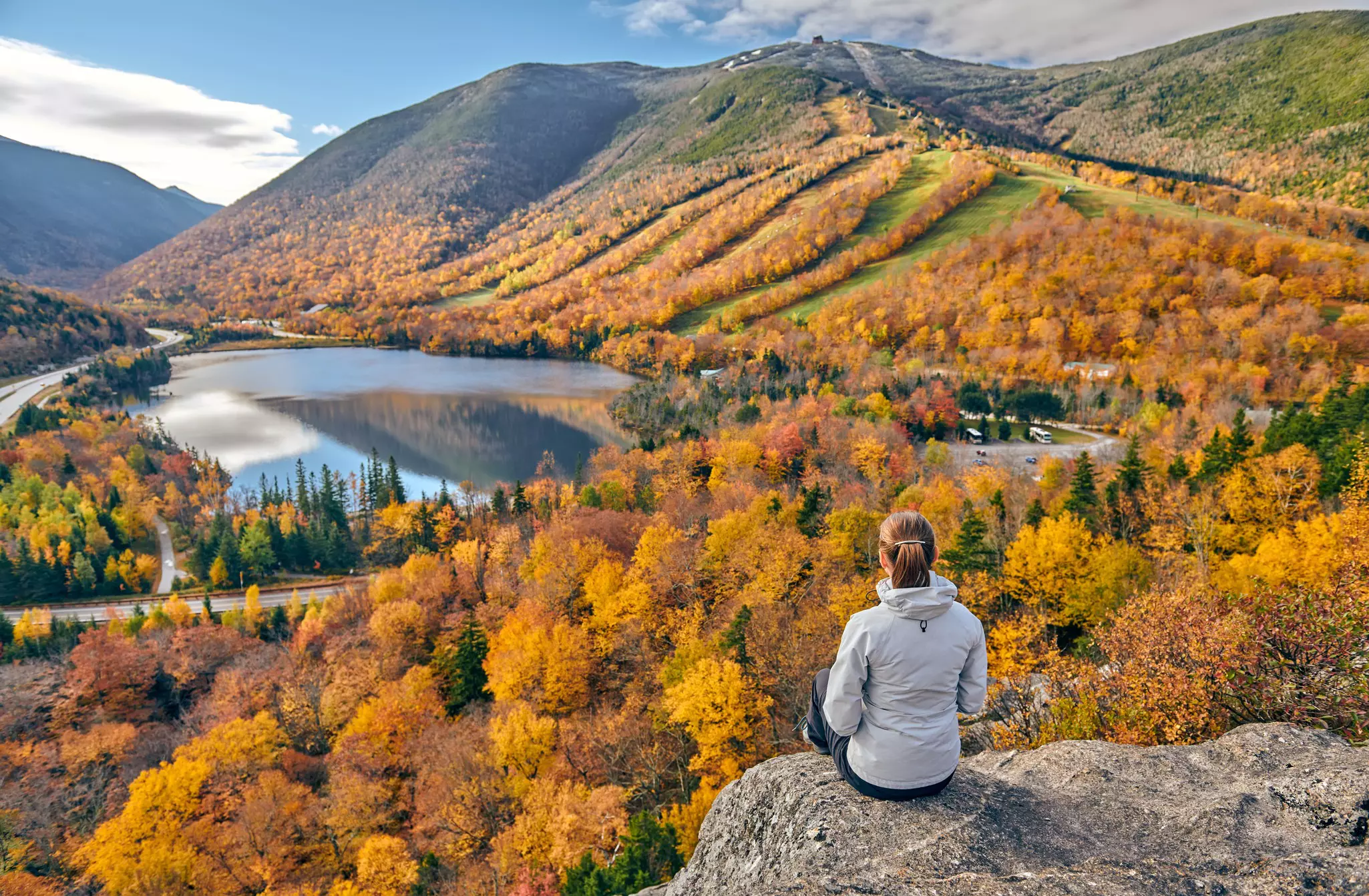 A woman sits on a rock at a viewpoint looking down over woodland in the golden colors of fall.