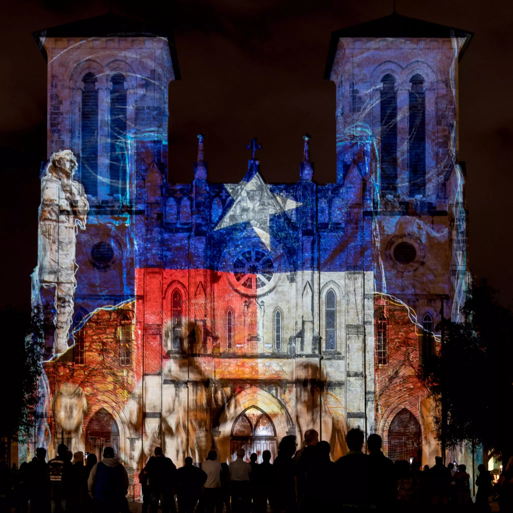 People are silhouetted against the facade of a cathedral in San Antonio, Texas, with a projection on the building.