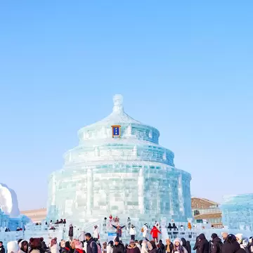 Crowds in heavy coats on packed snow before a round, tiered building carved from blocks of ice under a clear blue sky.