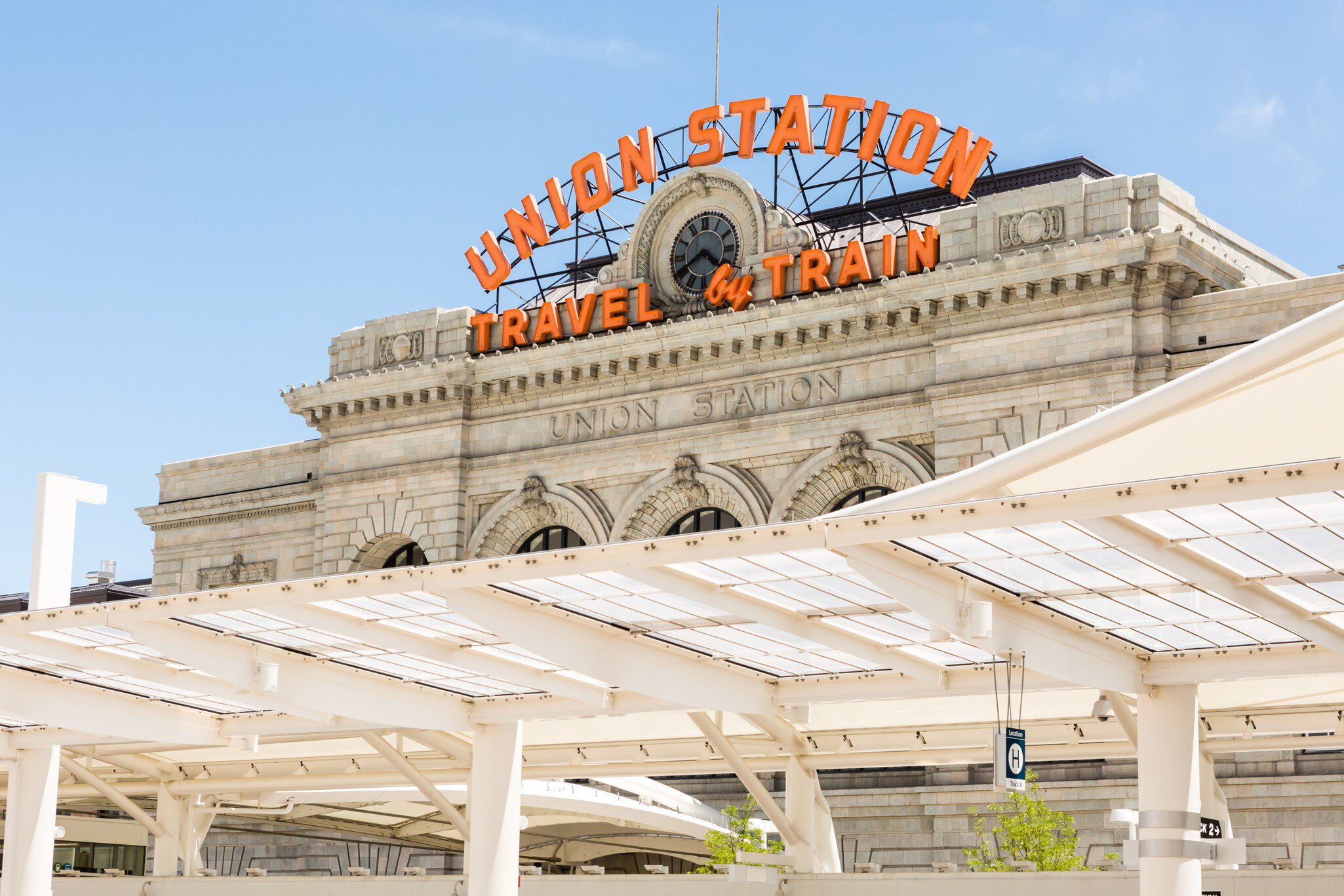 Orange neon sign UNION STATION TRAVEL by TRAIN above a clock on grey stone; white platform canopies below.