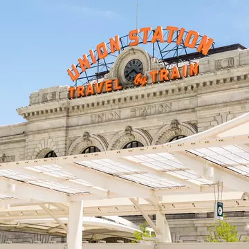 Orange neon sign UNION STATION TRAVEL by TRAIN above a clock on grey stone; white platform canopies below.