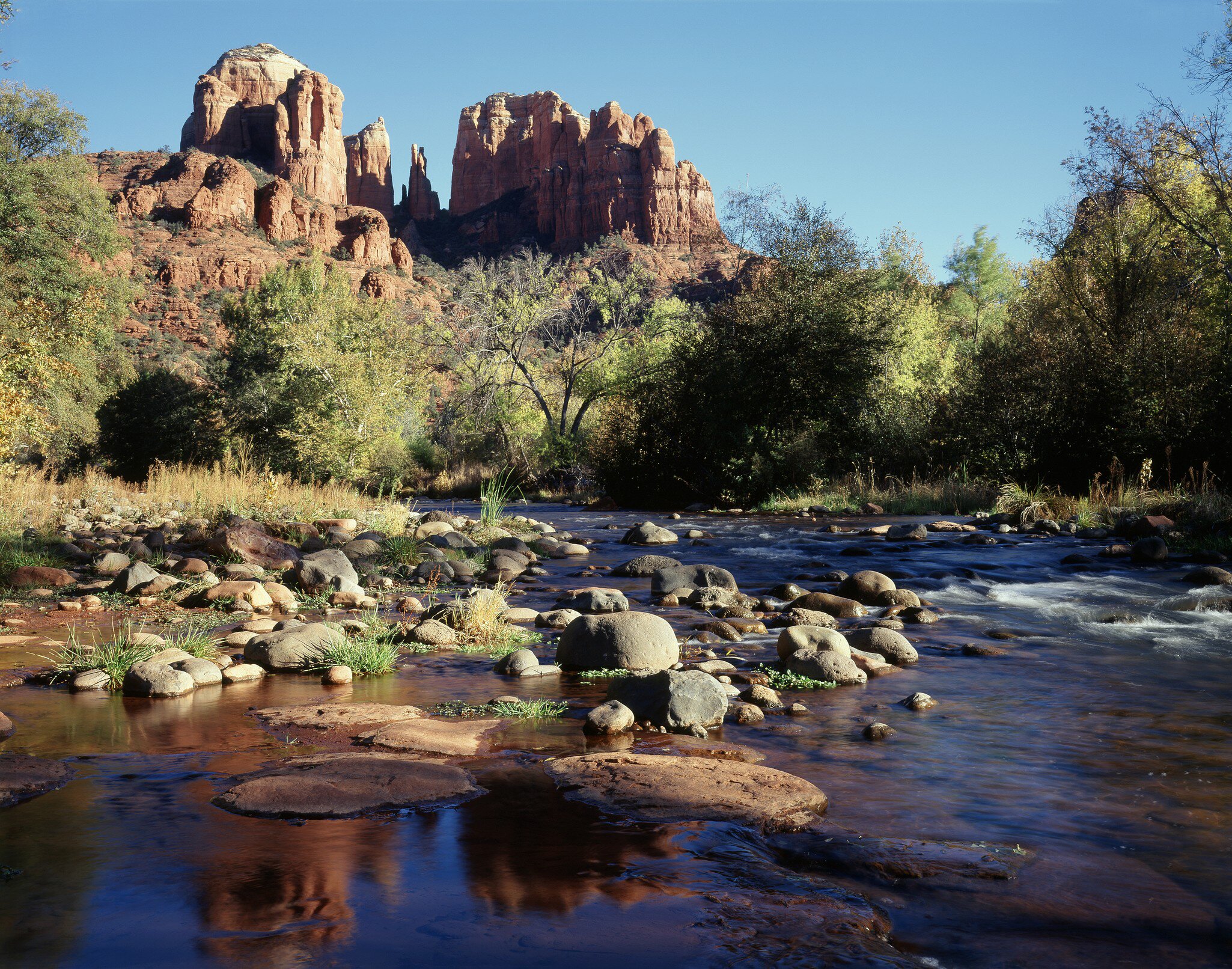 Shallow creek runs over rounded boulders and flat red slabs, with autumn-tinged trees under a bare blue sky.