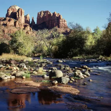 Shallow creek runs over rounded boulders and flat red slabs, with autumn-tinged trees under a bare blue sky.