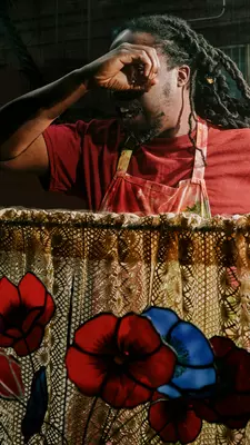 A man in a red shirt peers into a window half covered with a lace curtain and in front of a glass flower arrangement.