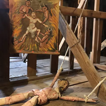 A wooden marionette without a costume is displayed on a wooden shelf. A small painting hangs behind the puppet.