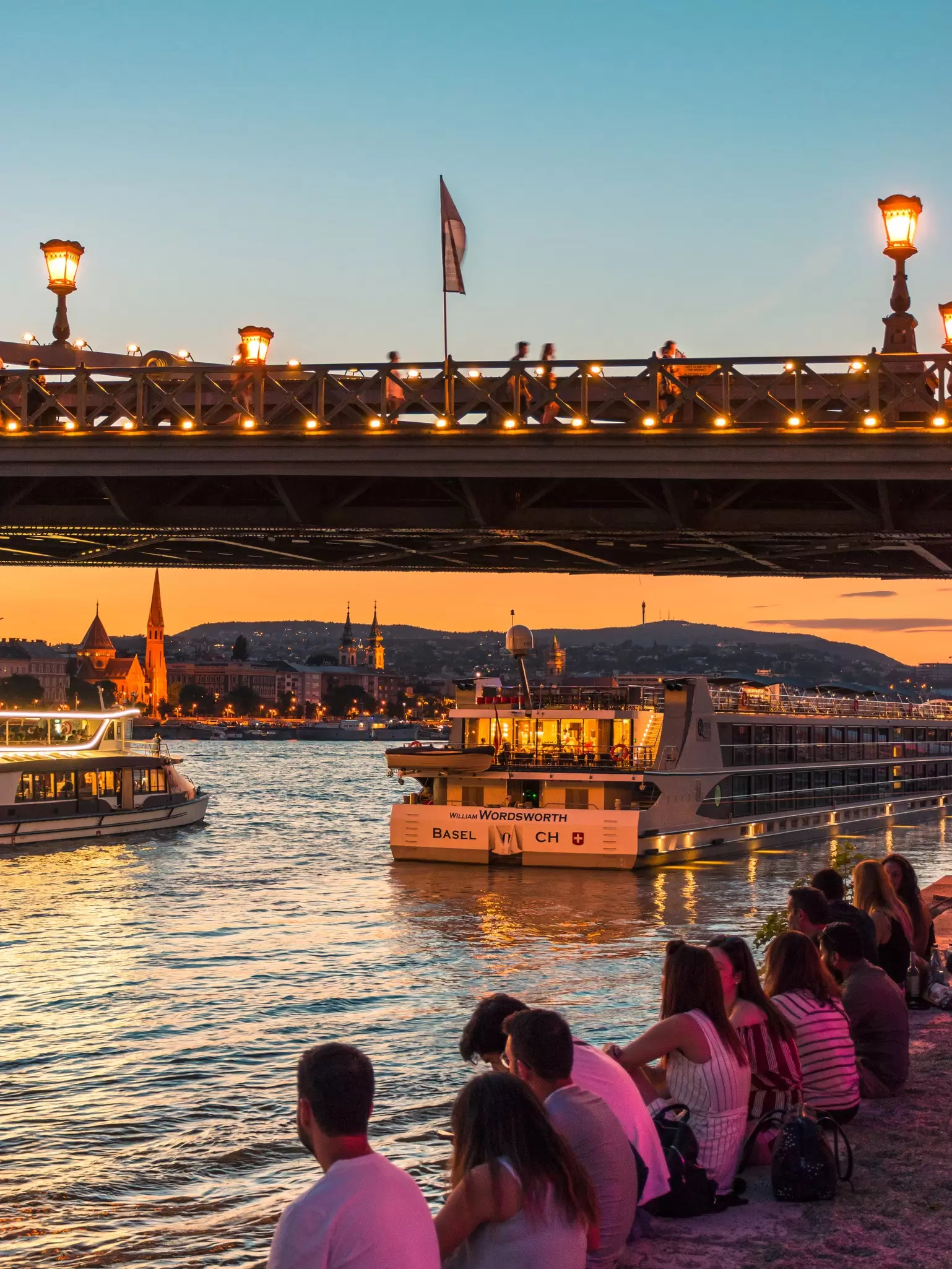 People watching the sunset on the shore of the Danube next to the Széchenyi Chain Bridge in Budapest, Hungary.