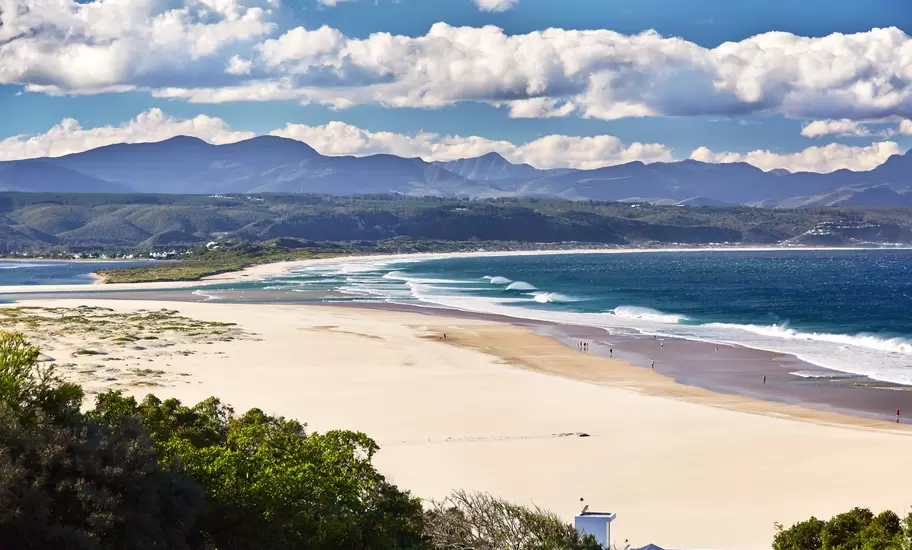 Long white sand beach curving round Plettenberg Bay, with breaking waves, a lagoon and mountains behind under cumulus clouds