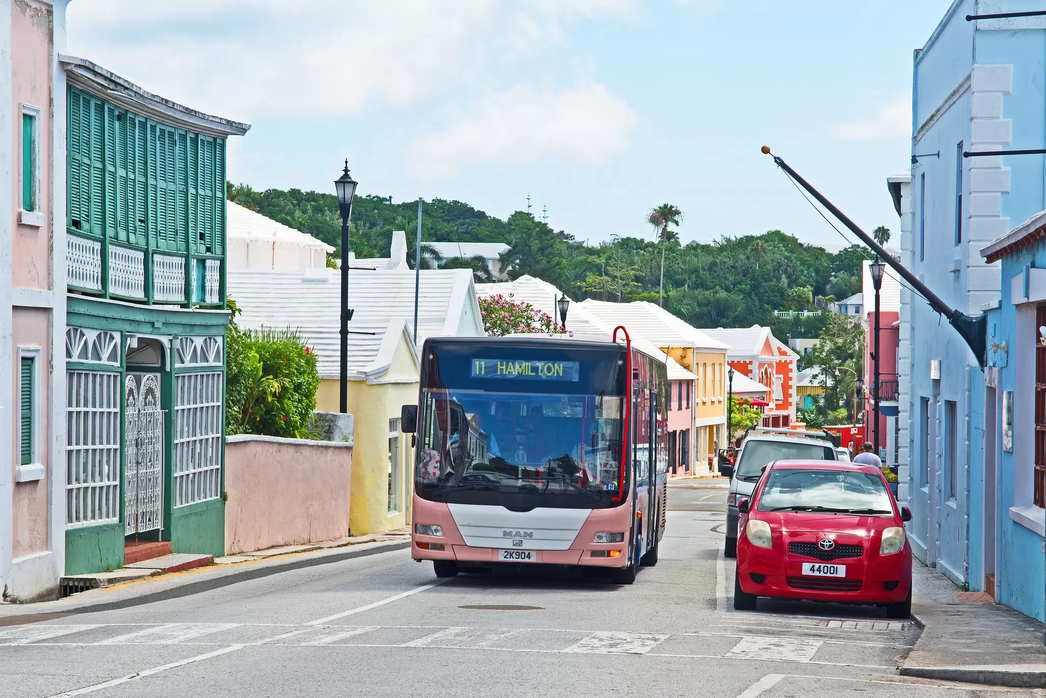 A pink bus driving past parked cars on a street lined with colorful buildings