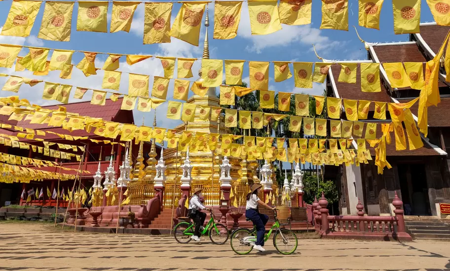 Two people on bicycles pedal past the gold stupa of a temple. Numerous yellow flags hanging from strings flutter overhead.