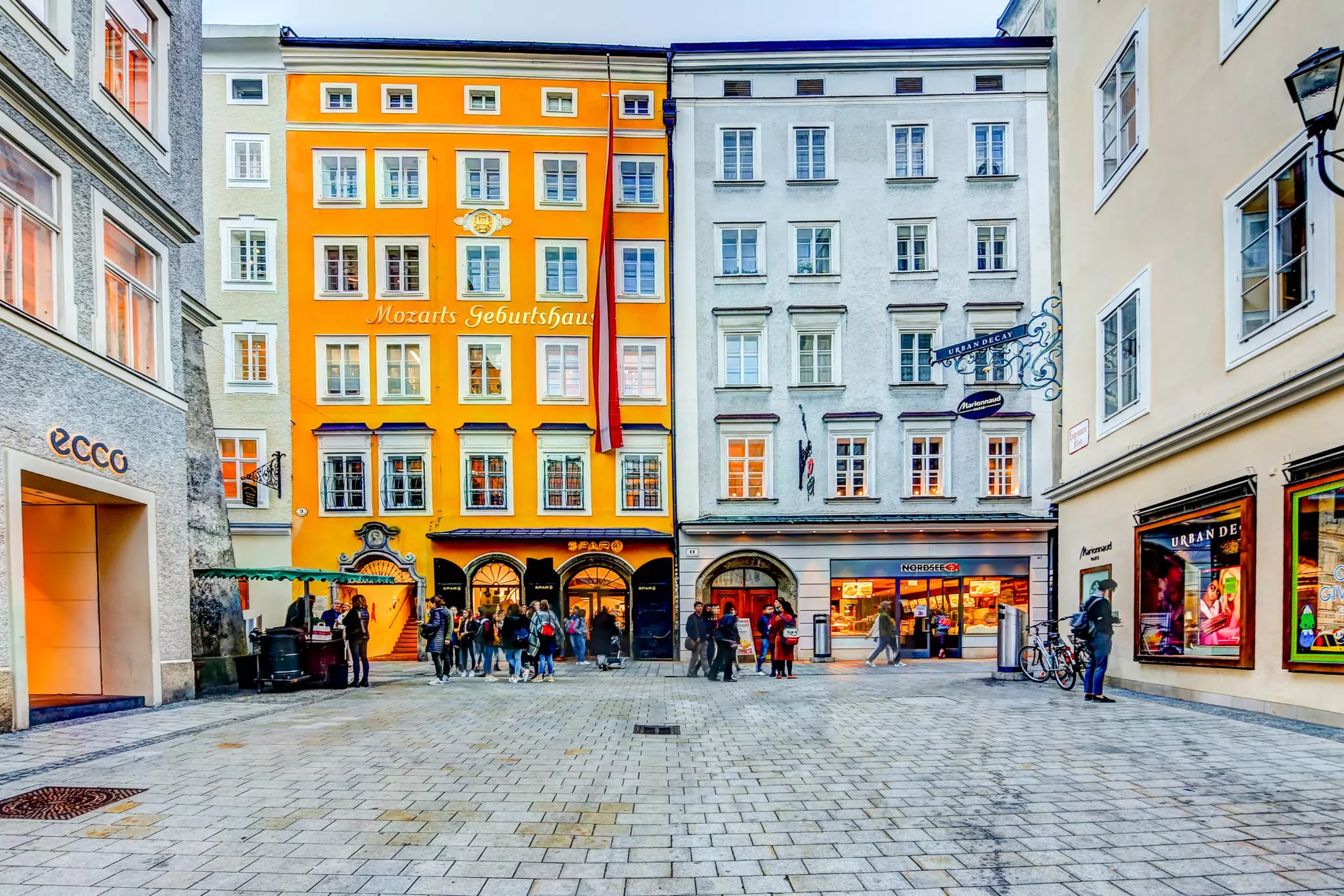 Plaza of concrete pavers with historic buildings in front, one yellow and one gray, and commercial shops to the right and left.
