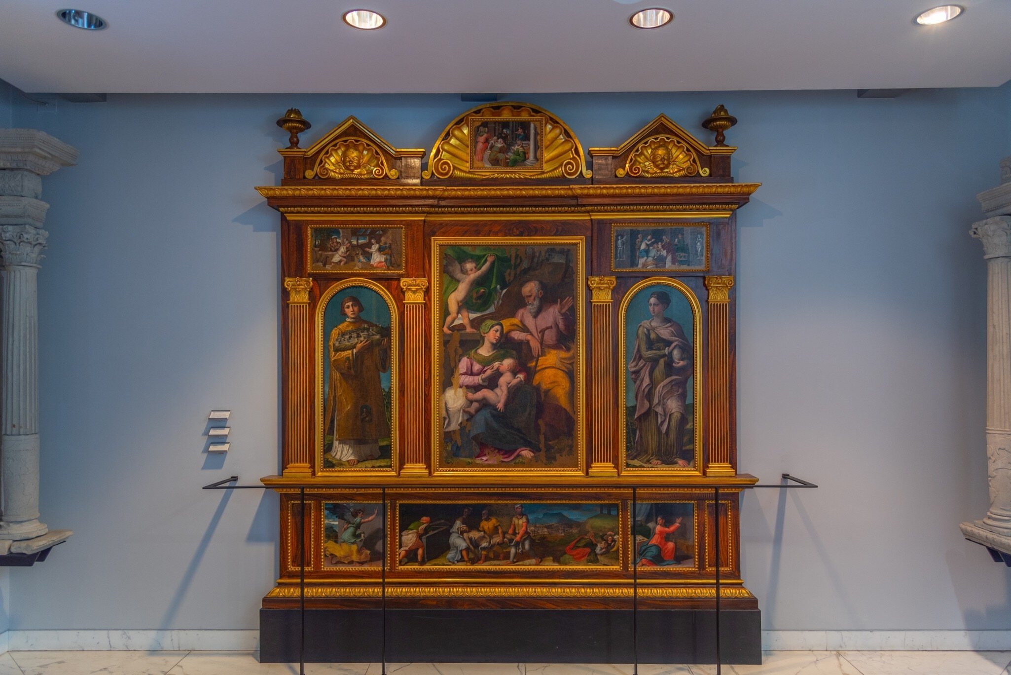 A gilded Renaissance altarpiece of the Virgin and saints on a pale blue wall.