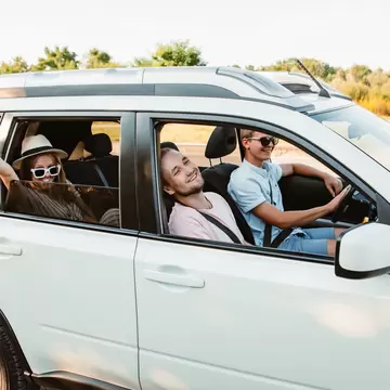 Smiling friends wave from the open windows of a white SUV on a sunny road trip.