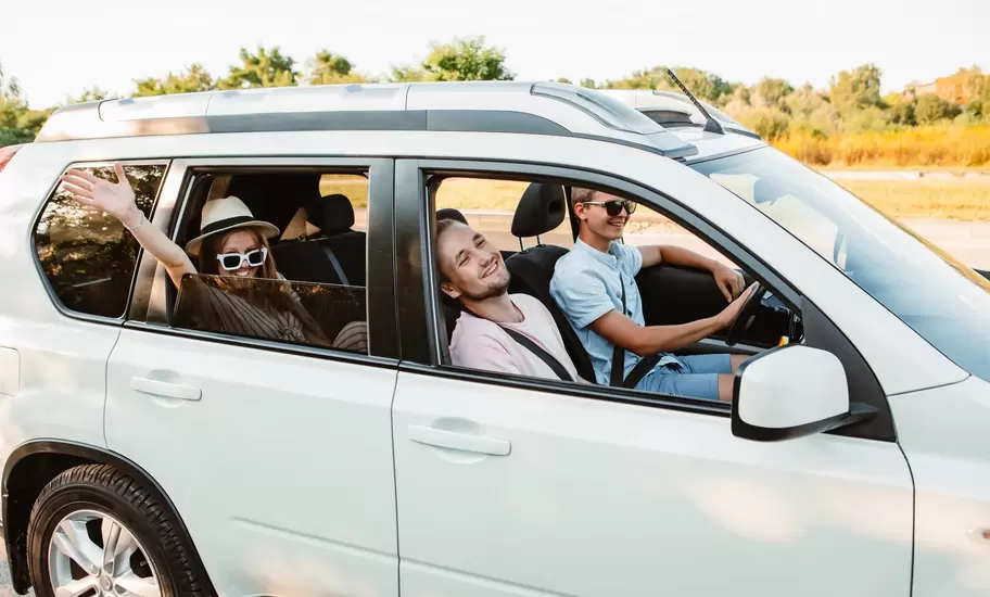 Smiling friends wave from the open windows of a white SUV on a sunny road trip.