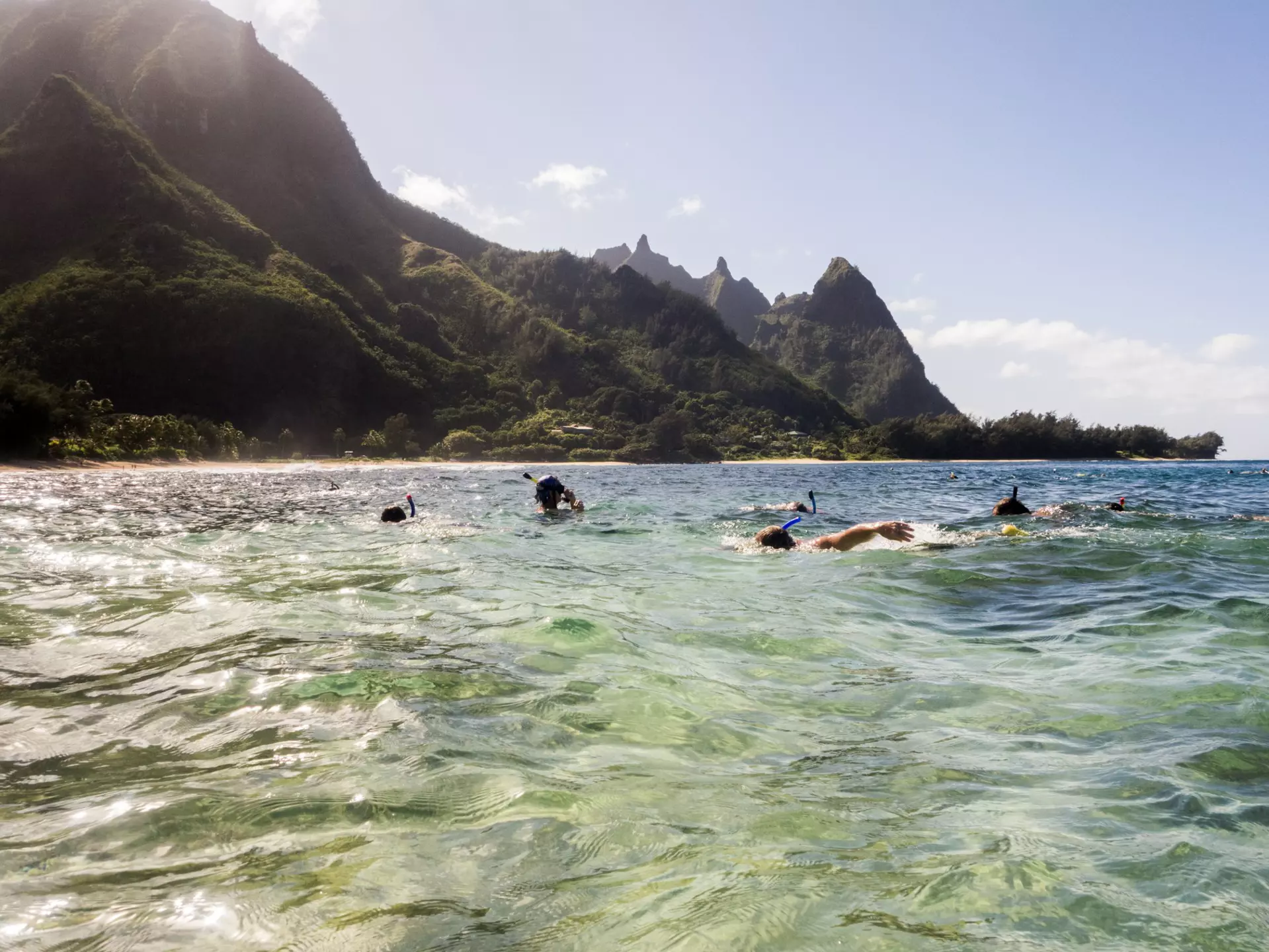 A group of people scuba diving and snorkeling off the coast of Kaua‘i on a sunny day with dramatic cliffs in the distance