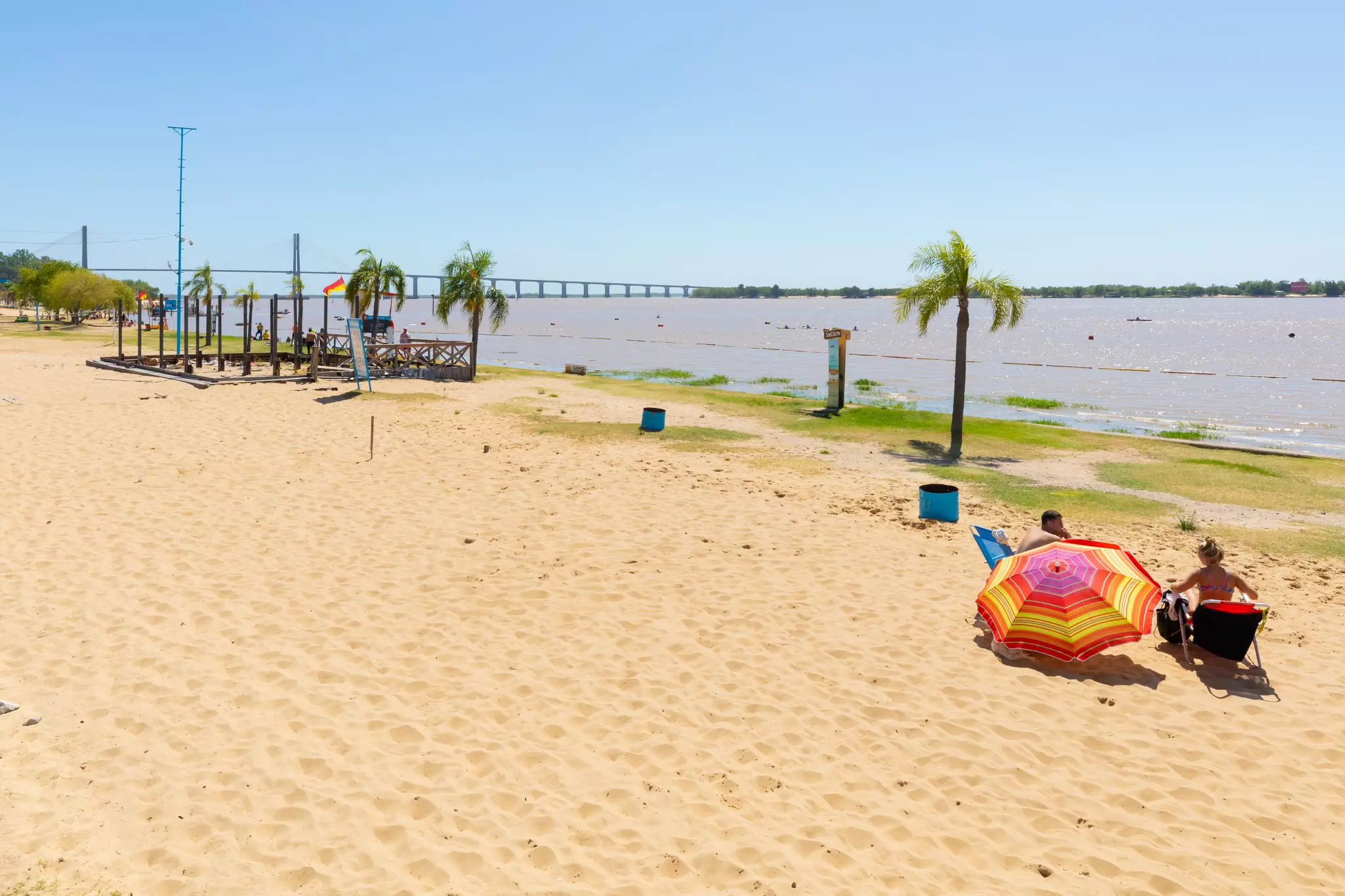 People sit under a colorful sunshade on the sandy shores of a riverside beach.