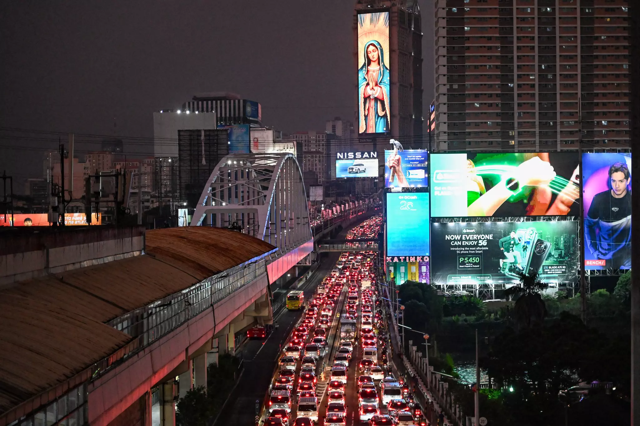 An aerial view of the rear brake lights of vehicles stuck in traffic on a six-lane highway. Illuminated billboards and an arched bridge are visible to the sides of the highway.