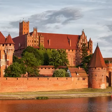 Long red-brick fortress with steep tiled roofs, a square tower and two conical gate towers above a calm river