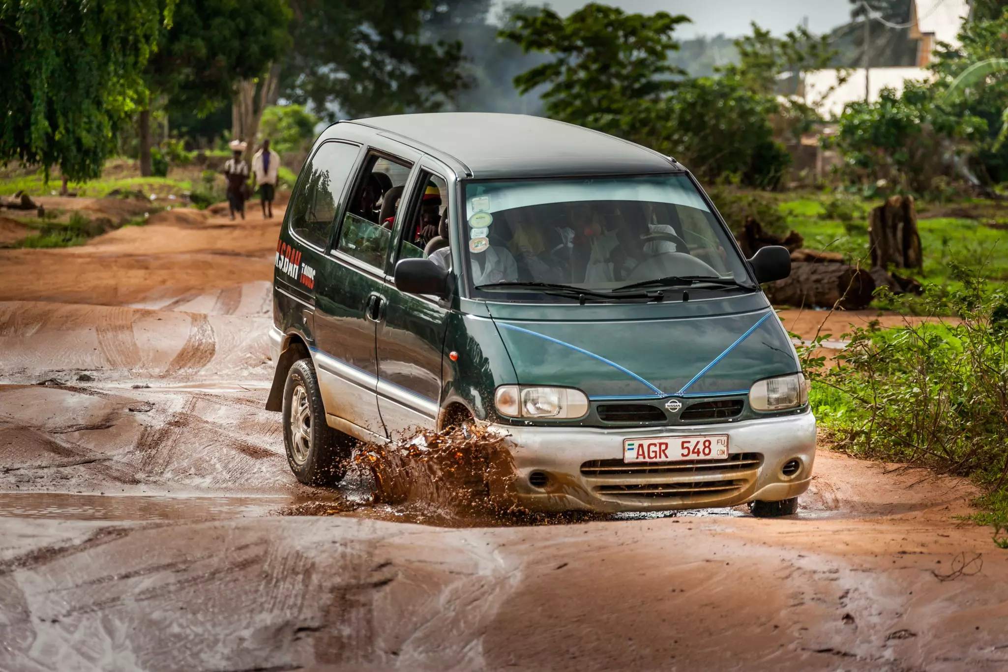A green minivan drives through a muddy puddle on an unpaved road.