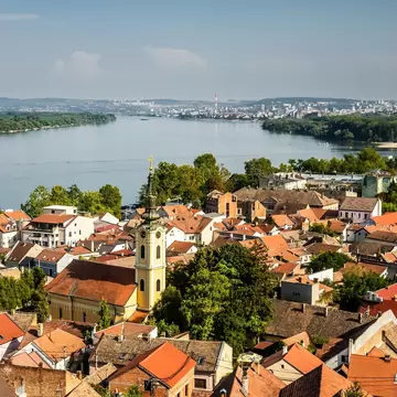 Yellow church with a tall clock tower and gilded cross amid orange tiled roofs, a broad river and distant city beyond