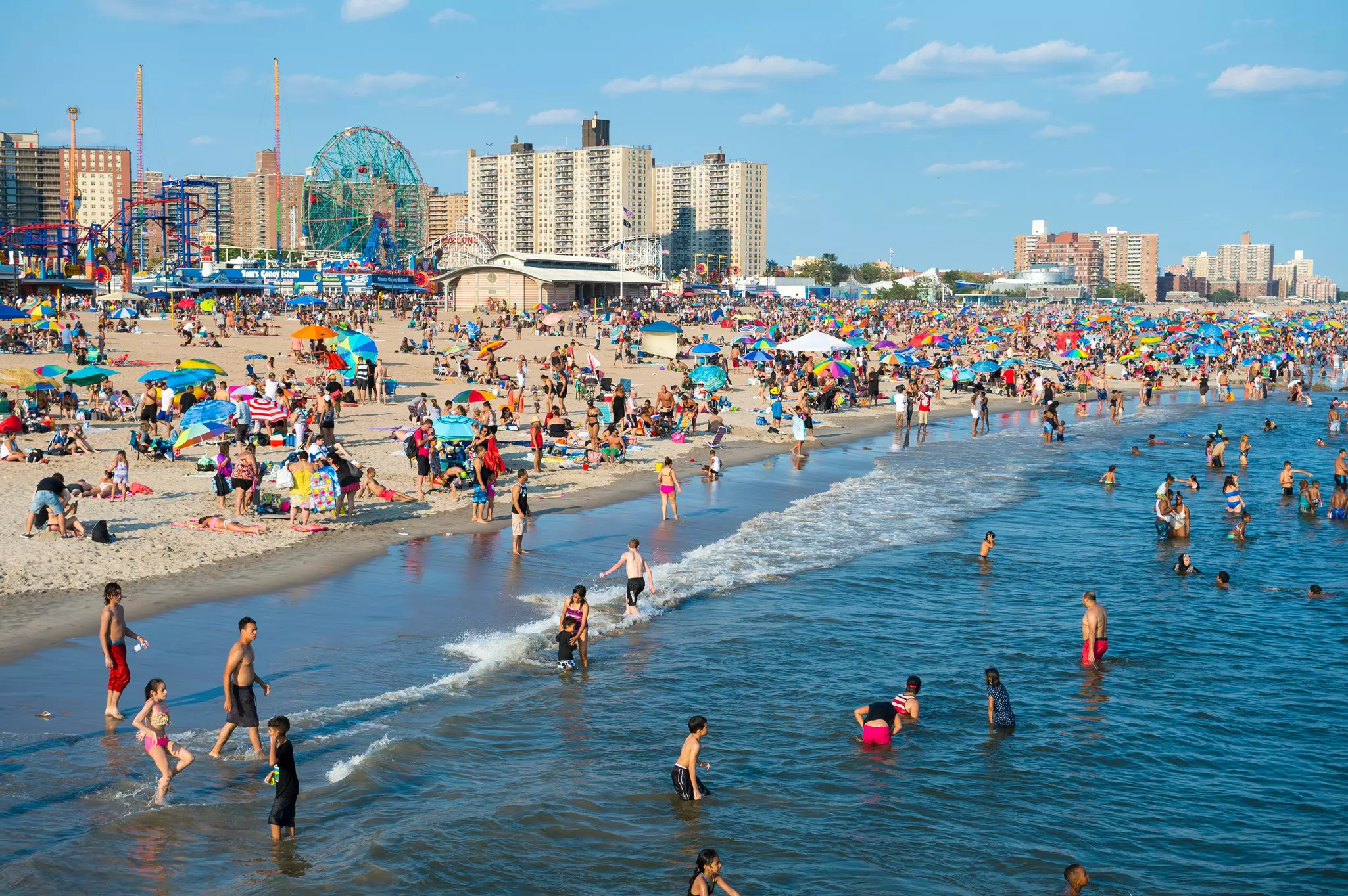 A view of people wading and swimming in the water at a city beach, its sand crowded with other people, and apartment towers and amusements visible in the distance.