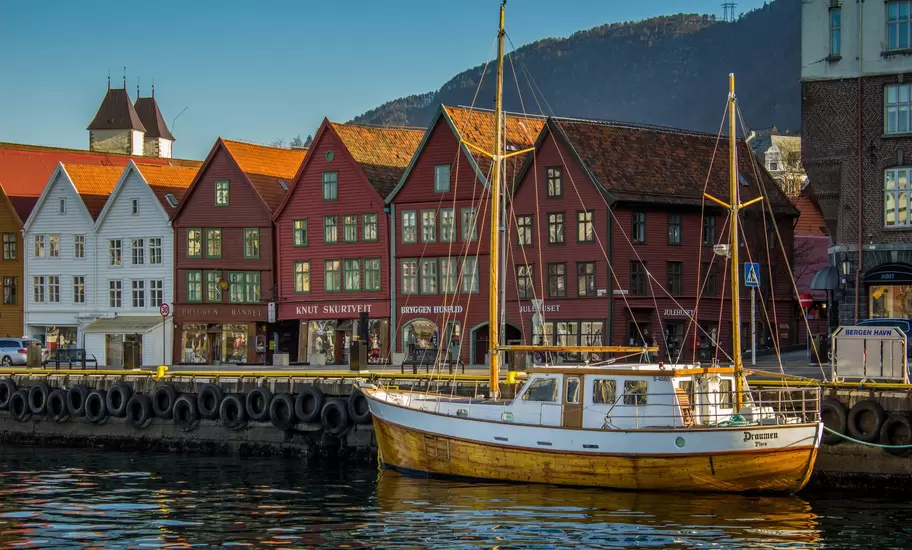 Bryggen wharf in Bergen: red and white wooden gabled houses behind a moored two-masted boat, wooded hill behind
