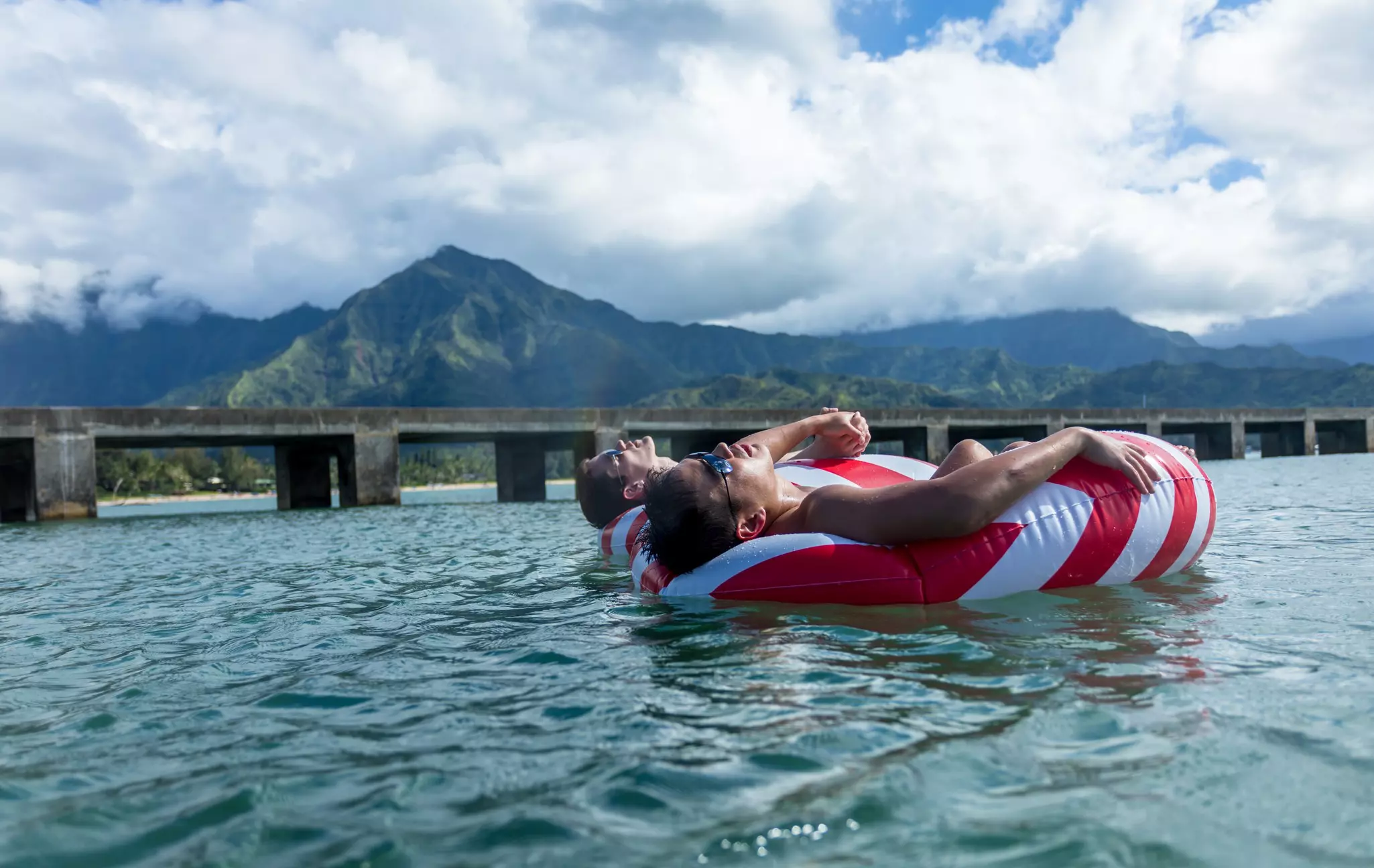 Two men holding hands while floating on red-and-white rafts in the water in Hawaii