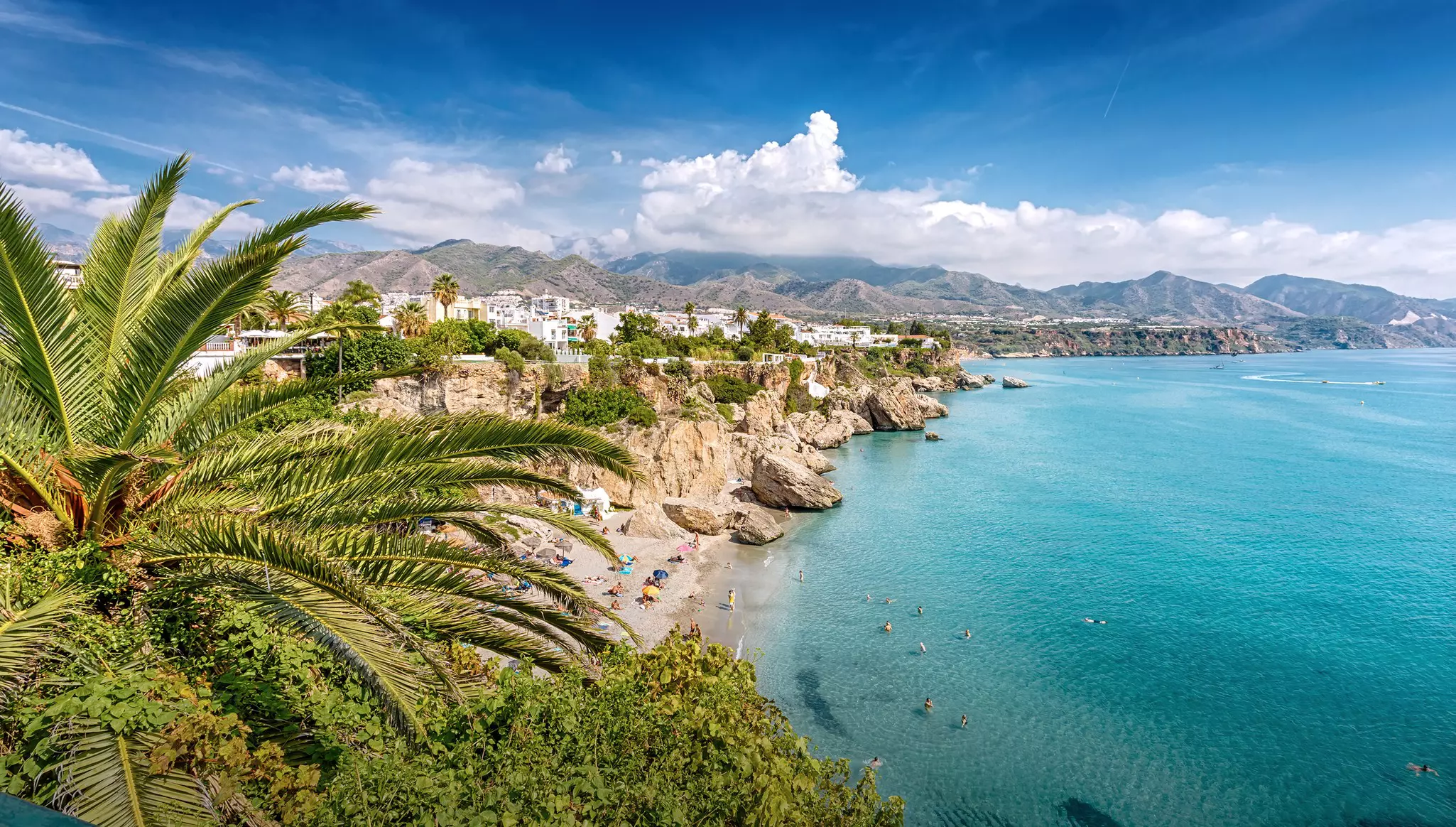 Aerial shot of sandy beach with large rock outcroppings and people swiming in clear blue sea with a line of houses in the distance and low mountains further beyond.