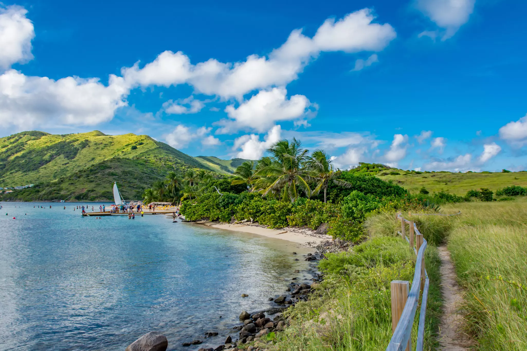 Lush vegetation grows along the shore of a tropical island. A pier with swimmers and a sailboat is visible in the distance.