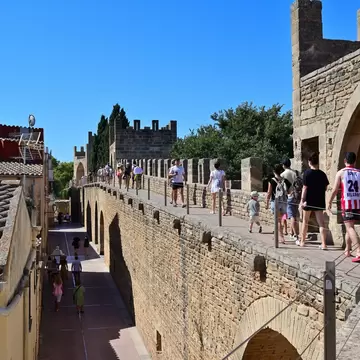 People walking a stone wall-walk between battlemented towers under blue sky, a narrow street and tiled roofs below.