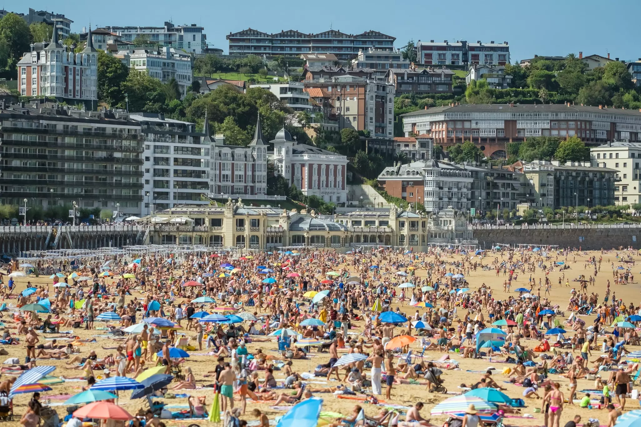 Thousands of people crowd on the sand of a city beach. Hotels and apartment buildings line the beachfront boulevard.