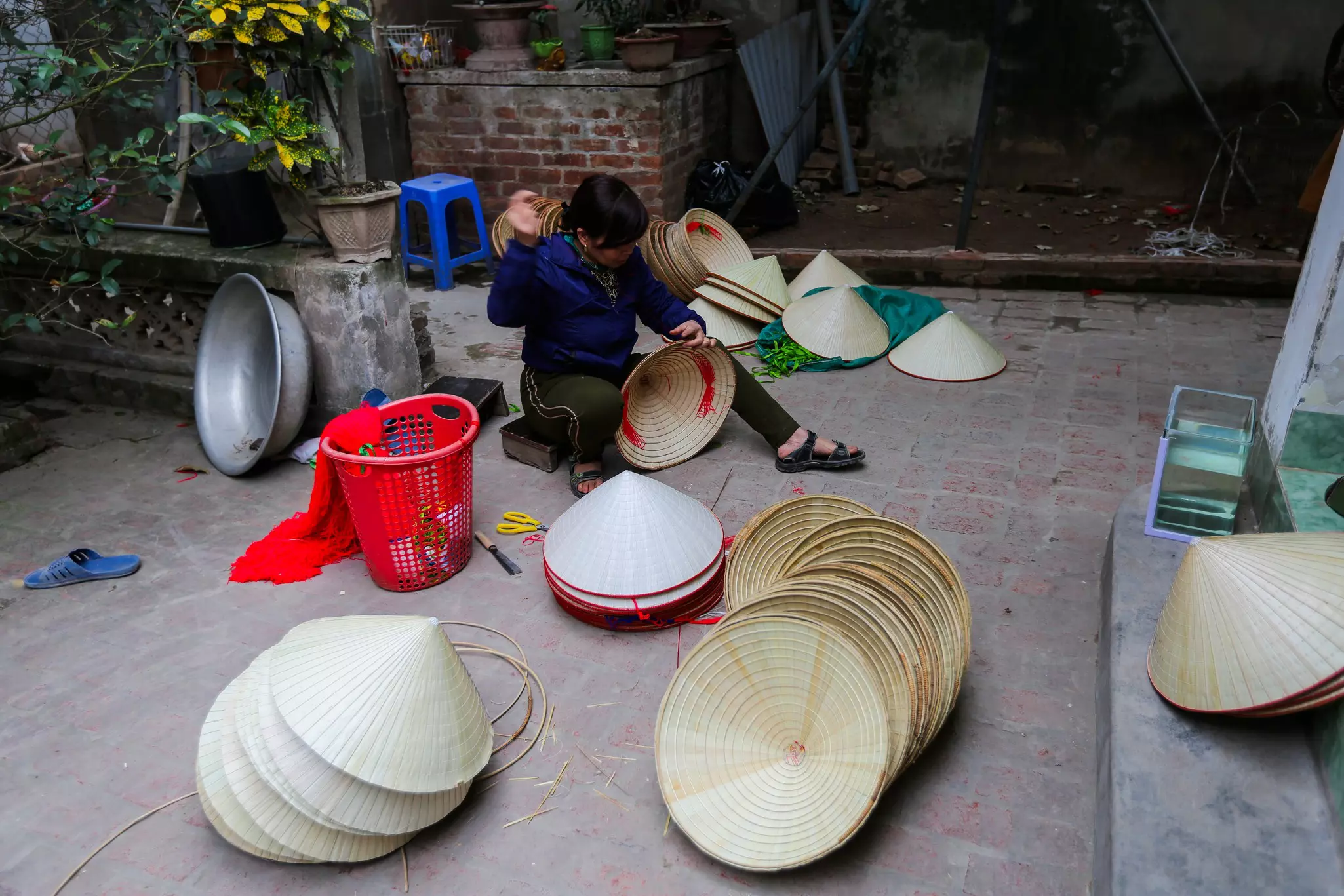 A woman sits on a very low stool while making a traditional conical hat. More hats in various stages of creation are piled around her.