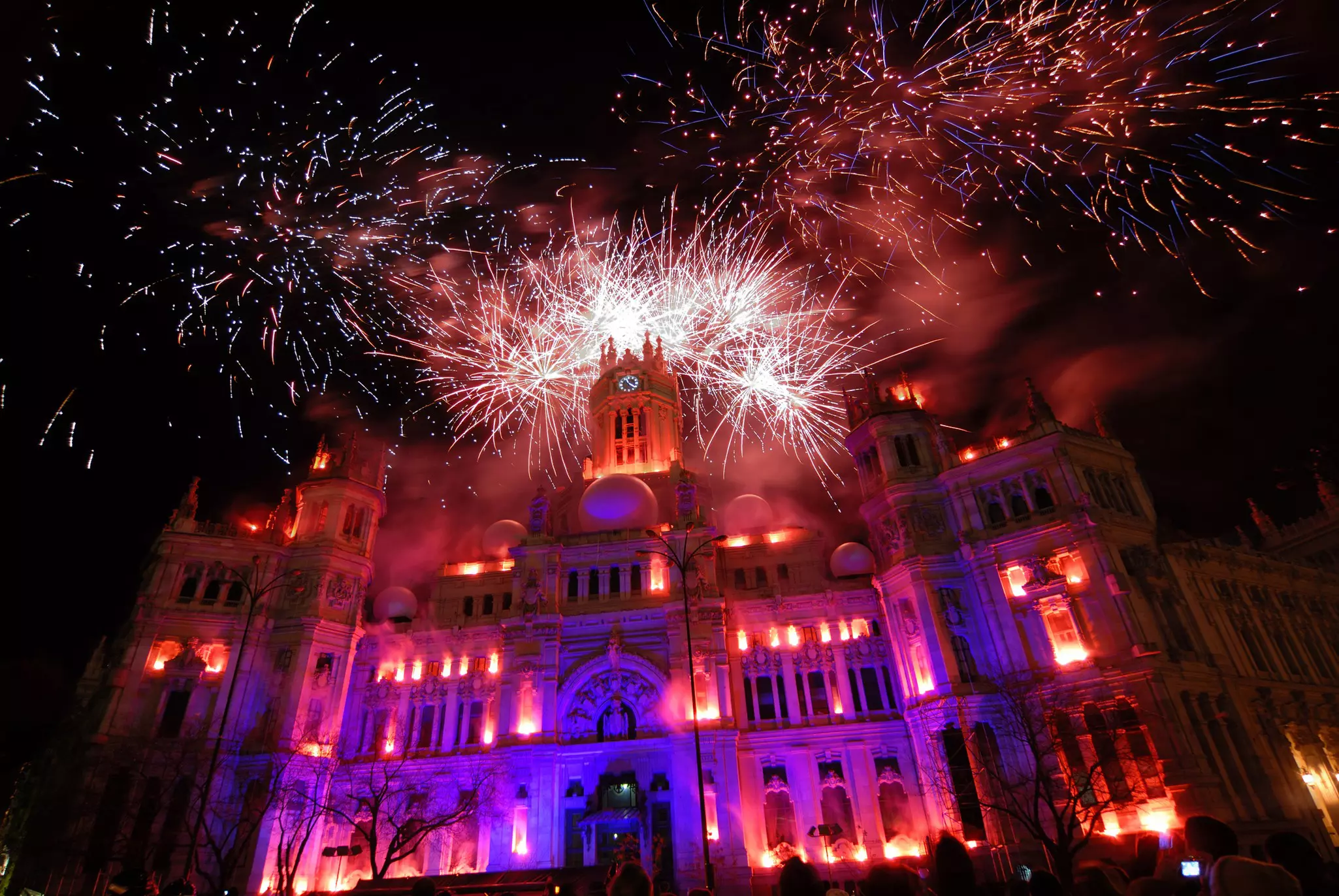 Fireworks light up behind a large building whose windows are aglow with additional lights at night