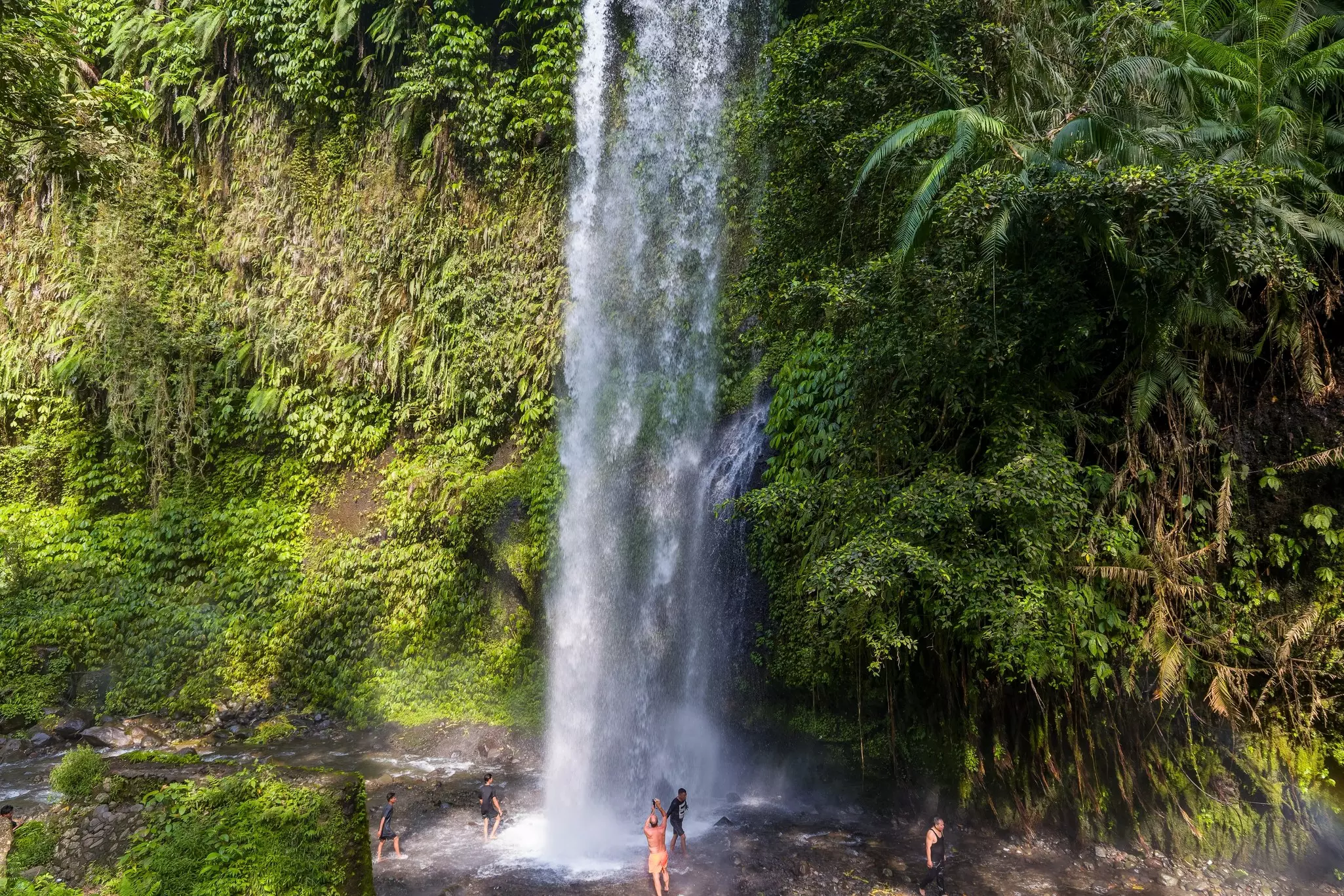 People stand in a pool below a vast cascading waterfall in Lombok, Indonesia.