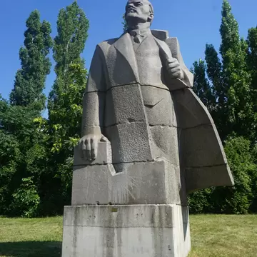 A monumental grey stone figure of Lenin in a flared greatcoat on a concrete plinth, standing on mown grass against poplars and blue sky.