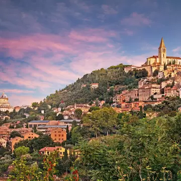 Hillside town of terracotta buildings topped by a Gothic church, with a domed church to the left, under a pink evening sky.