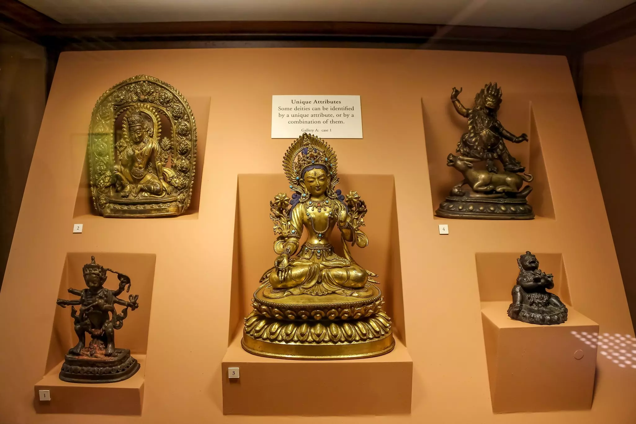 A bronze Buddha surrounded by Hindu gods figurines on orange background in a museum display