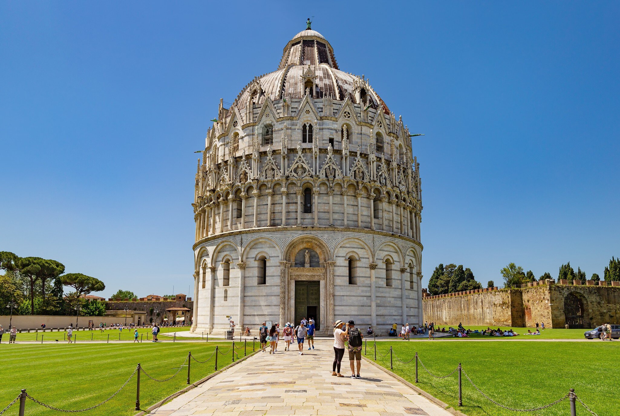 Visitors on a path leading to the domed marble Baptistery on the Piazza dei Miracoli in Pisa, with city walls at right