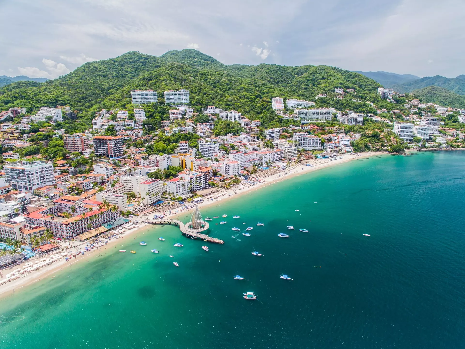 Boats dot the beachfront at Puerto Vallarta, Mexico, with hotels and green hills behind. 