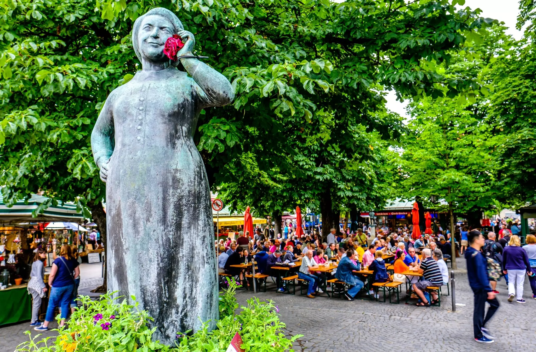 A statue stands among foliage. Behind tables are full of people dining from nearby market stalls