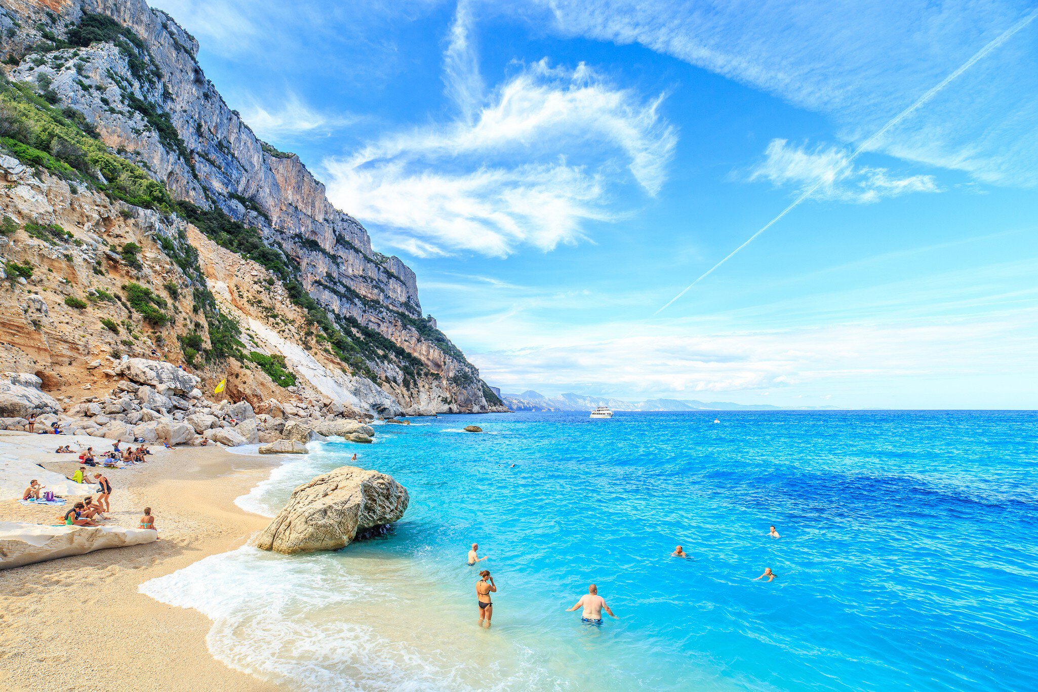 Swimmers in the blue ocean off a sandy beach backed by rocky cliffs on a sunny warm day.