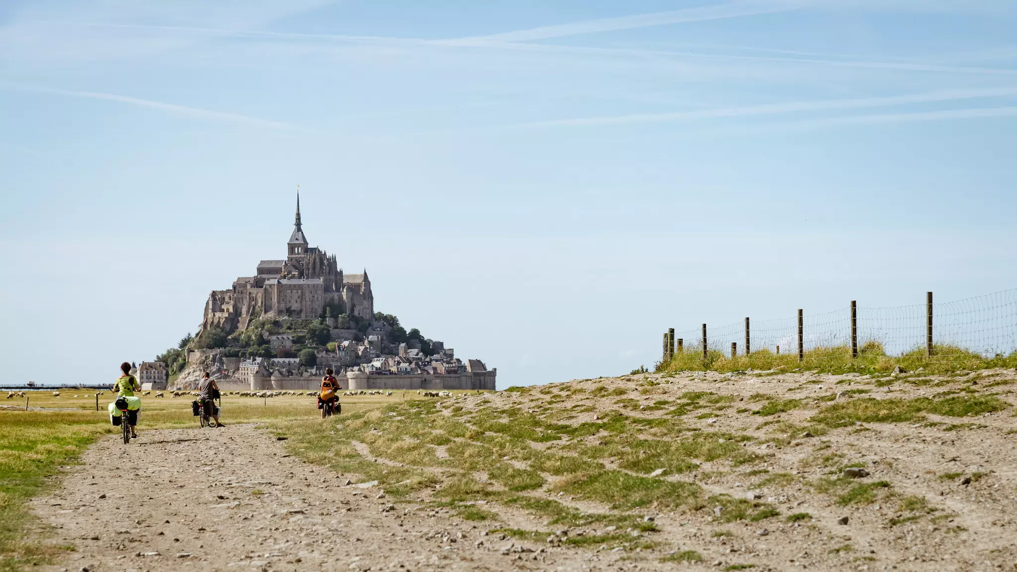 Three cyclists with loaded packs on their bikes pedal along a path towards a raised island with a monastery on the top of it.