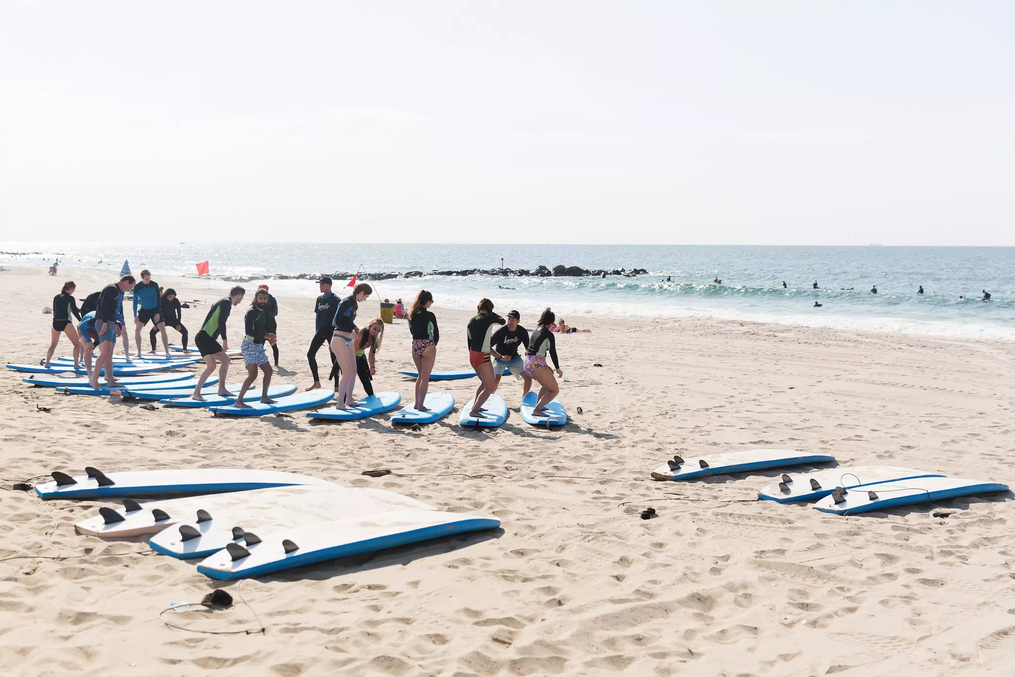 A group of people stand on surf boards on a sandy beach while receiving a lesson from an instructor.