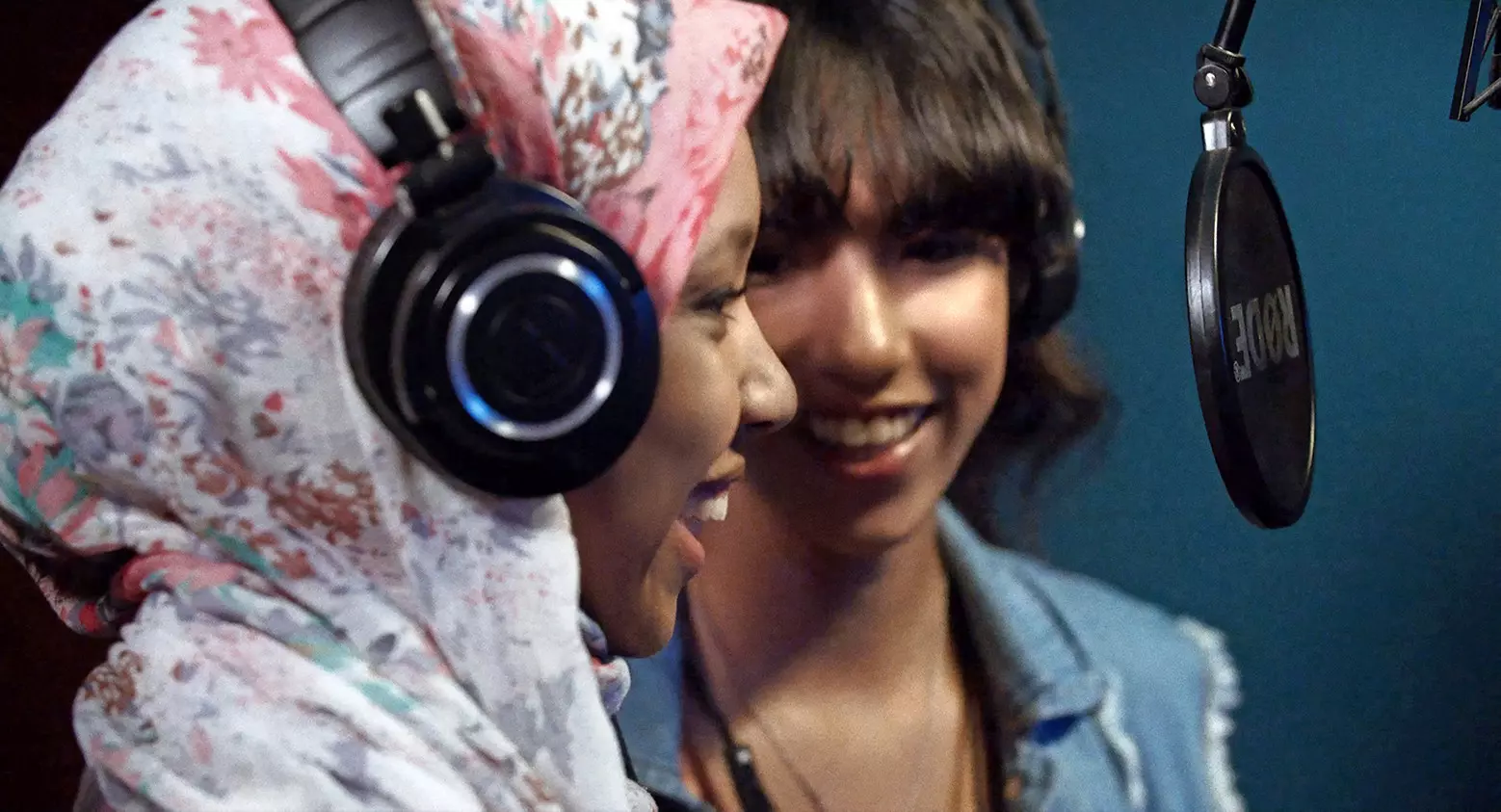 Two women, one wearing a pink patterned headscarf and headphones, speak into a hanging microphone.