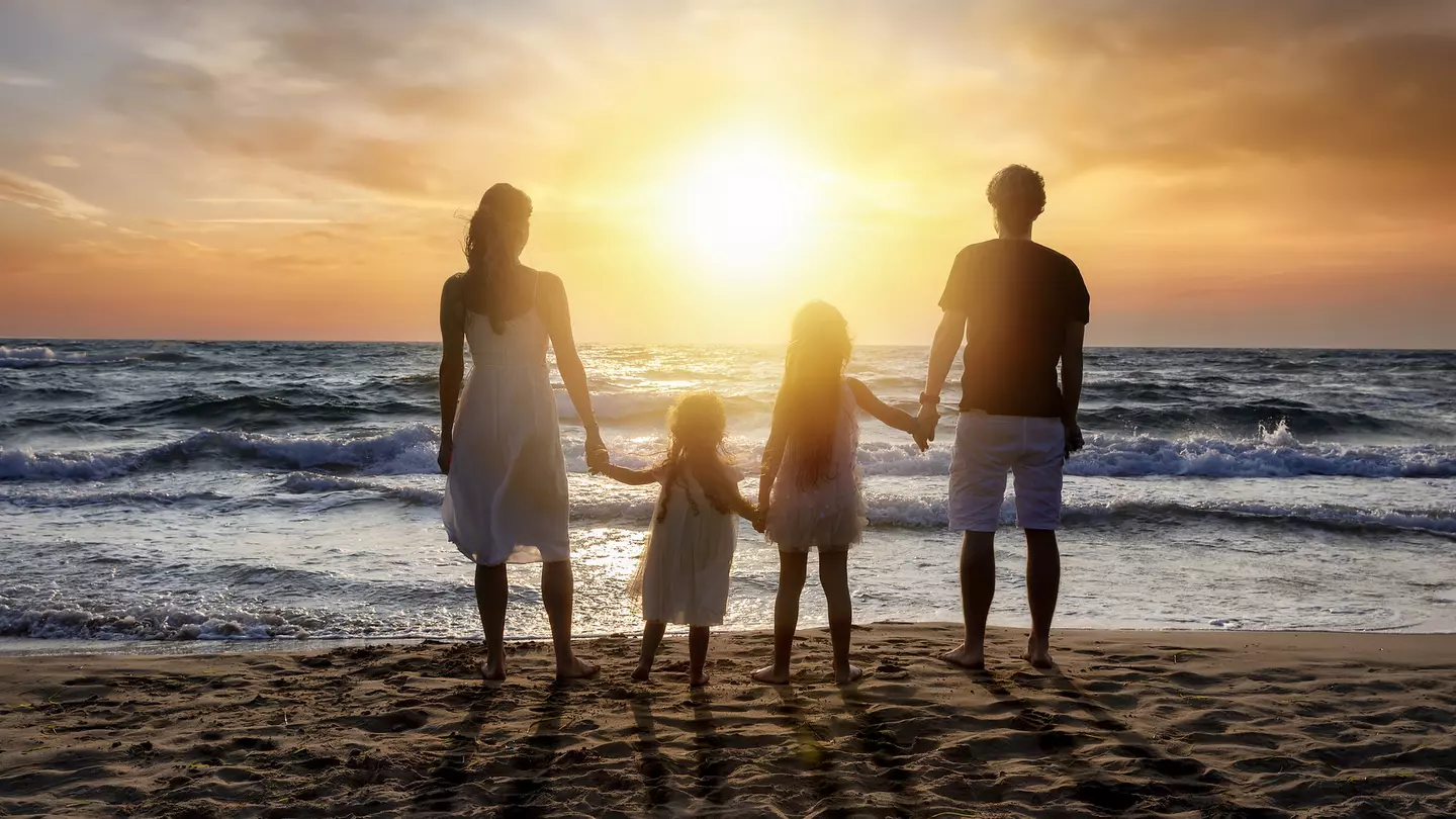 Rear view of parents holding hands with daughters standing at beach against orange sky